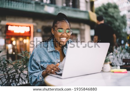 Joyful dark skinned female blogger typing text of publication using laptop computer working remotely, cheerful African American woman in spectacles share positive content and media files on netbook
