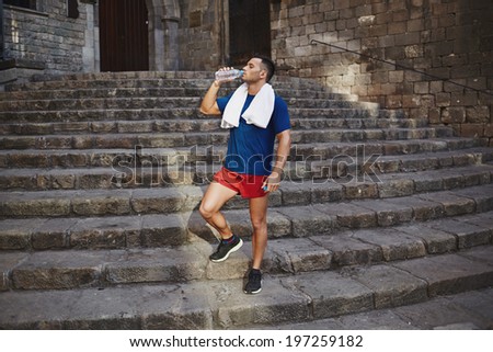 Male runner with a towel around his shoulders drinks water after a tiring morning jog