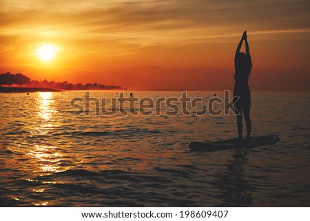 Sup yoga performed by beautiful girl on the background of the bright orange sunset, yoga training on the beach