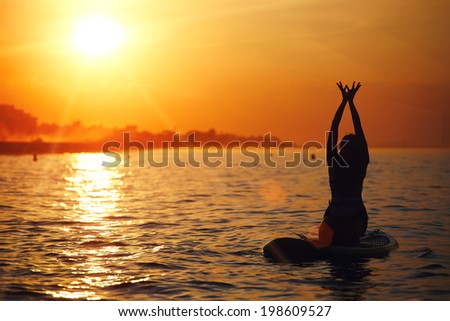 Evening yoga meditation on the paddle surf board at amazing bright sunset reflected on the ocean, silhouette of the girl engage yoga