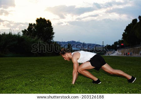Athletic muscular runner dressed in the white t-shirt and black shorts ready to start standing on the grass in the beautiful park, sky background, health lifestyle and fitness concept