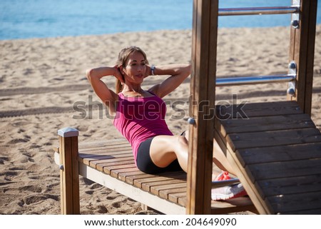 Attractive middle age woman doing abdominal crunches on the beach, female runner doing workout on beautiful sea background, fitness and healthy lifestyle concept