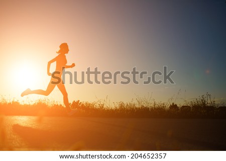 Silhouette of athletic girl running down the beautiful road at amazing orange sunset, female runner with muscular body at evening run,  fitness and healthy lifestyle concept