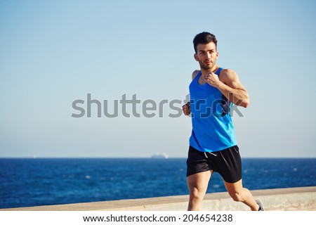 Attractive male runner with muscular body running along the sea on beautiful sky background, athletic sportsman on the evening run, fitness and healthy lifestyle concept