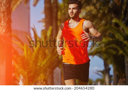 Muscular athletic man running on the jogging track on the palm trees background, male runner on the evening jog, fitness and healthy lifestyle concept