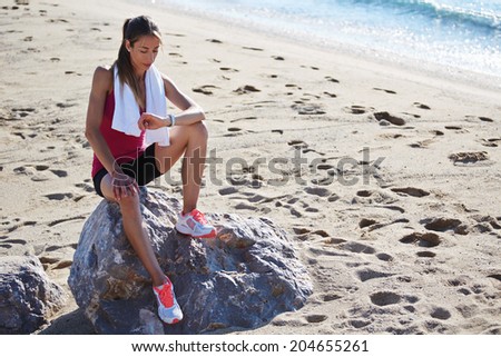 Beautiful female runner in bright sportswear with towel on the shoulders looking the time in her wrist watch, female runner resting on the beach after intensive run, fitness and healthy concept