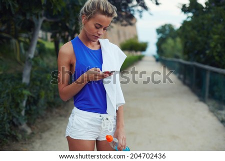 Beautiful sporty blond girl holding telephone in the hands and looking to the screen, evening run in the park, fitness and healthy lifestyle concept