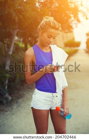 Beautiful female runner holding telephone in the hands and looking to the screen, evening run at sunset, fitness and healthy lifestyle concept