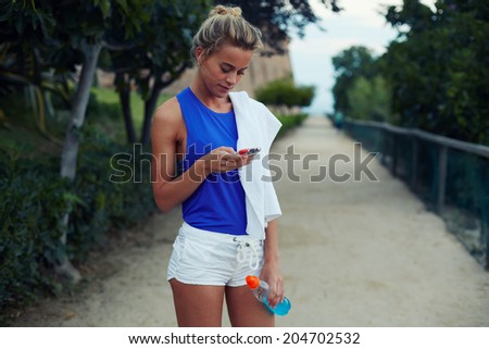 Blond sporty girl with athletic figure resting in the park and looking to the telephone screen, run in the beautiful park, fitness and healthy lifestyle concept