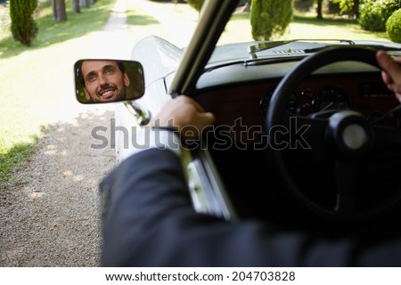Handsome successful man looking to the car mirror and smiling, stylish rich man driving luxury car on beautiful green road, lifestyle and successful business concept
