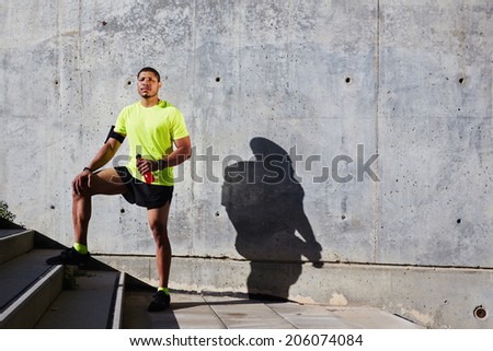Attractive runner with strong muscular body resting after run, african american sportsman in bright sportswear standing on concrete wall background at sunset, fitness and healthy lifestyle concept