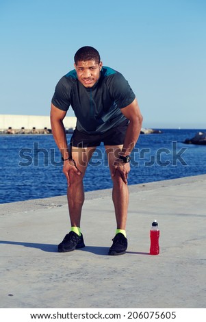 Young muscular build runner resting tired after training on beautiful sea port background, attractive male runner raking break after evening running outdoors, fitness and healthy lifestyle concept