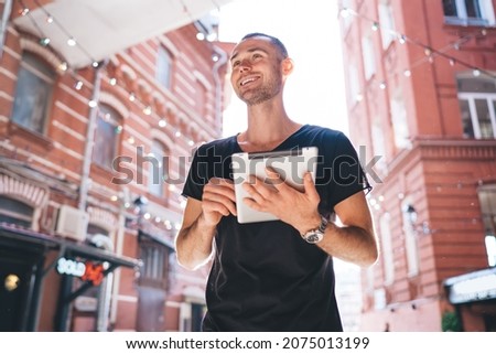 Below view of cheerful hipster guy in casual T-shirt using touch pad technology and smiling outdoors, joyful Caucasian male tourist enjoying vacations time holding digital tablet or modern ebook Imagine de stoc © 