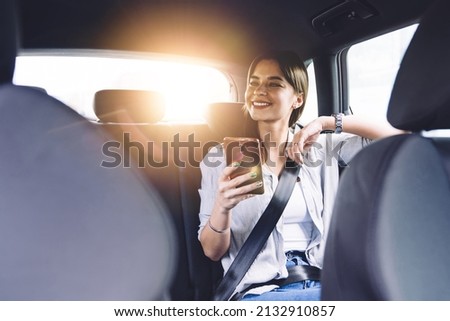 Cheerful positive young female in casual wear sitting in automobile backseat with seatbelt fastened and looking away while using mobile phone Stock foto © 