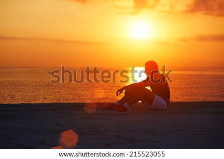 Tired after morning jogging athletic runner resting on the beach, male runner taking break after training outdoors, runner resting on colorful sunrise background, healthy lifestyle concept