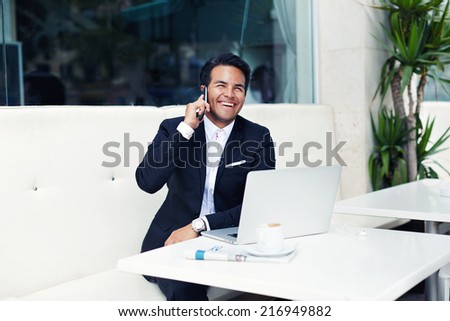 Attractive businessman sitting at desk in bright terrace of lounge bar cafe, handsome man talking on mobile phone and smiling, laughing wealthy man talking on mobile phone, man having a phone talk