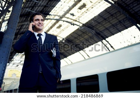 Businessman standing on railway platform using mobile phone, rich man in suit talking on the smart phone and smiling looking away, traveling businessman having mobile conversation on the way to work