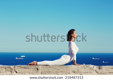 Beautiful woman in white clothes seated in yoga pose on blue sea background, healthy young woman meditating yoga enjoying amazing view of sea from high, practicing yoga at sunny day, feeling healthy