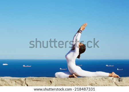 Beautiful brunette girl makes yoga pose on high altitude with sea and sky horizon on background, seated in one of yoga pose young woman with perfect body engage yoga meditation outdoors