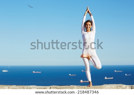 Attractive brunette girl smiling standing in yoga pose on beautiful sea background, healthy woman smiling during yoga training outdoors, happy woman practicing yoga enjoying sunny day at summer