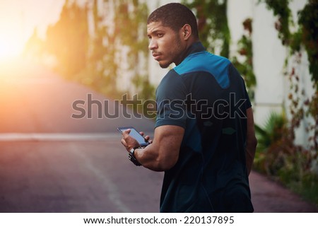 Attractive strong build runner resting after self training walking on the road, male runner using mobile phone, dark skinned jogger holding mobile smart phone walking outdoors