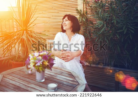 Beautiful woman enjoying sunny evening on house roof terrace in Barcelona,brunette middle-aged woman smiling seated on the terrace with beautiful plants and flowers, relaxation at summer sunny evening