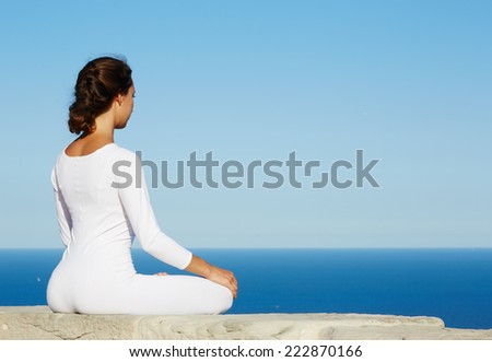 Woman seated in yoga pose on amazing ocean background, woman meditating yoga enjoying sunny evening, brunette woman makes yoga meditation sitting on mountain hill on amazing altitude, meditating woman