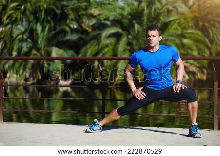 Beautiful male runner stretching before workout, fit man doing stretching exercises outdoors, young male sportsman stretching and preparing to run with beautiful green palm trees on background