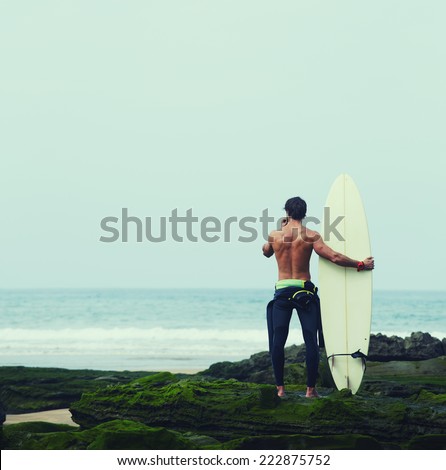 Young surfer holding surfboard while standing on the beach and looking at ocean to find the perfect spot to go surfing waves, professional surfer with surf board looking at ocean, filtered image