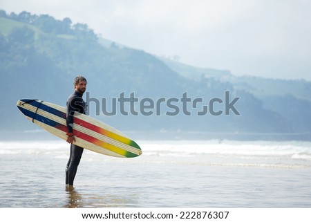 Young surfer standing agains the ocean looking the waves, professional surfer in black wetsuit holding with one hand big beautiful surfing board with multi-colored stripes, sunny day on ocean beach