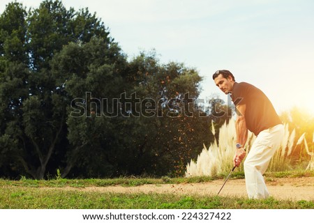 Professional male golf player hitting by driver standing on golf course at sunny evening, handsome man hitting golf ball from golf sand trap with sun light on background