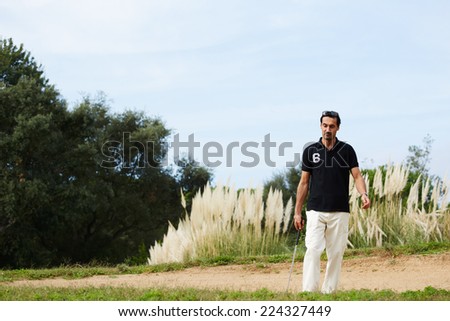 Handsome man at recreation, rich wealthy man in sport clothes holding golf driver walk on green golf field, confident and satisfied golfer holding driver while walking on golf course