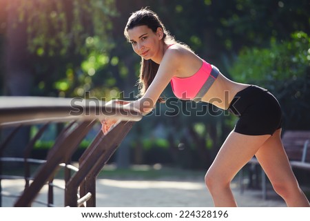 Young attractive female runner with perfect body resting in the park at sunny day, fit sexy girl taking a break balking on lake fencing in beautiful park, perfect sunny day for do sport outdoors