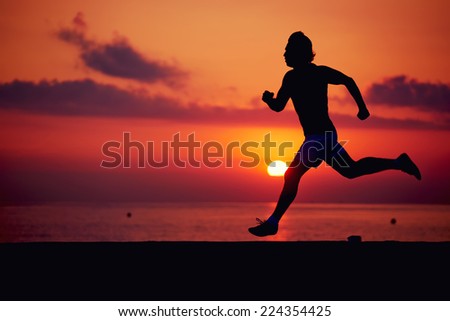 Silhouette of muscular build athlete running fast a log the beach, runner in action jogging against colorful sunrise over the sea, male jogger with muscular body in action