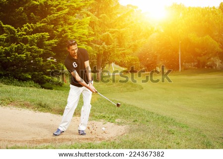 Strong golf shot of handsome player standing on golf course, golfer on sand trap hitting golf ball to the hole, golfer man hitting golf ball standing on beautiful green course at sunny evening