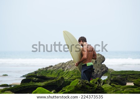 Beautiful brunette surfer walks on moss rocks carrying surfboard, sexy surfer man with muscular body going to surfing on big waves