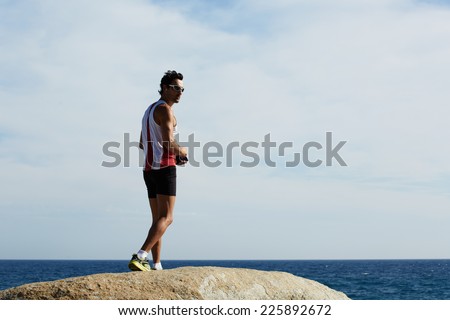 Young athlete listening to music during workout training outdoors, strong fitness man standing on big sea rock while resting after run, male jogger taking breach after workout training on the beach