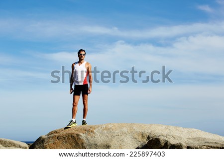 Exhausted fit runner after workout resting on seaside, successful winner standing on big hill, athlete standing on sea rocks at sunny summer day while resting after intensive training outdoors
