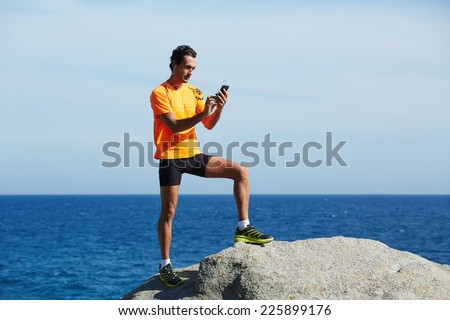 Athlete standing on sea rocks at sunny summer day while using mobile smart phone, male runner in fluorescent-shirt using mobile phone during workout training outdoors, jogger on sea horizon background