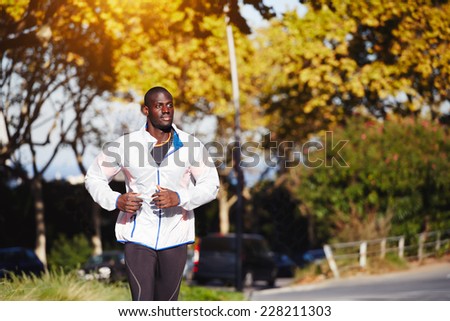 Young fit man in white windbreaker running at orange sunset outdoors, autumn running, attractive dark skinned runner jogging fast on beautiful orange leaves background in the park, sport concept
