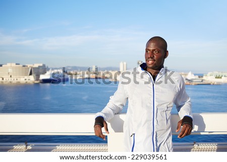 Attractive male runner in white wind breaker taking break standing on sea background, black fit man resting after workout outdoors, tired to run dark skinned runner resting after fitness training