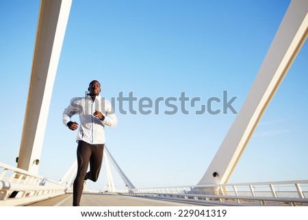 Attractive fit man running fast along big modern bridge at sunset light, black man doing workout outdoors, athletic runner in windbreaker jogging over bridge road with blue sky on background