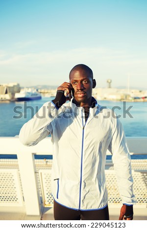 Attractive runner using mobile phone after fitness training, dark skinned sportsman with cell phone standing on beautiful sea port background