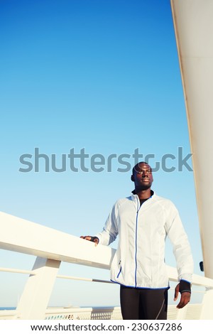 Handsome fit man dressed in windbreaker taking leaning on bridge fence taking break after workout, male runner resting after run standing on beautiful sky background, evening fitness training outdoors