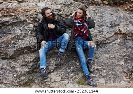 Beautiful couple of hipsters smiling sitting on mountain rock, friendly smile of two guys during adventure mountain expedition, fashionable couple having fun sitting outdoors