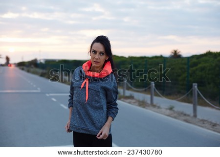 Fitness woman dressed in bi color hoodies standing on the road at sunset, attractive female runner taking a break after intensive run outdoors