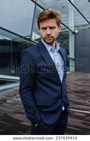Portrait of sad businessman standing with hands in suit pockets, desperate businessman standing against skyscraper office building, portrait of defeat businessman looking to the camera