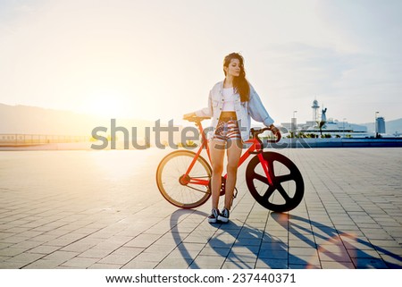 Attractive young woman win fixed gear bike posing outdoors with soft sunset light on background, pretty young brown haired woman standing with her modern pink bicycle at sunset, stylish hipster woman