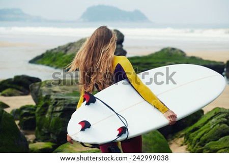 Blond long-haired surfer girl walking to the ocean holding her surfboard, female surfer holding copy-space surfboard