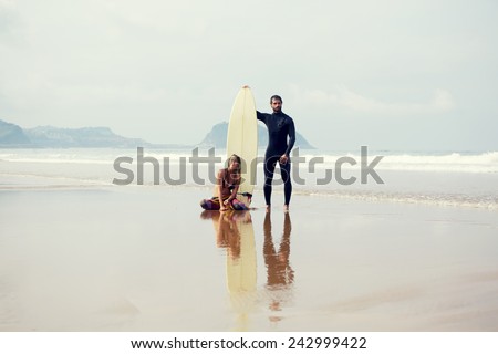 Beautiful surfer couple standing on the beach before their sunday surf lesson, young cute couple of surfers standing on amazing ocean and mountains background